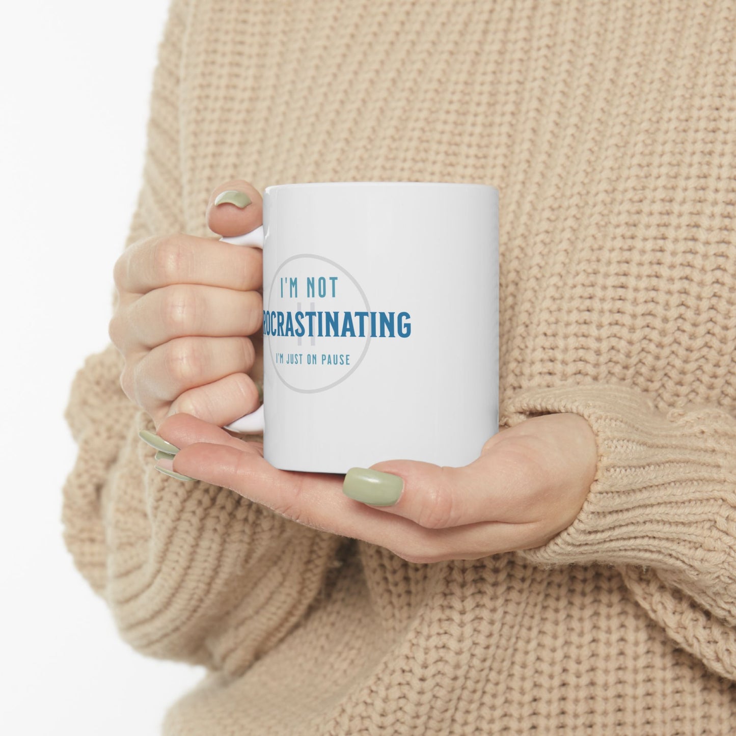 A person wearing a beige knitted sweater holds a Printify Procrastination Ceramic Coffee Mug featuring the IM NOT PROCRASTINATING, IM JUST ON PAUSE design. Their light green nail polish glints in the light, making it ideal for coffee lovers and quirky drinkware fans.
