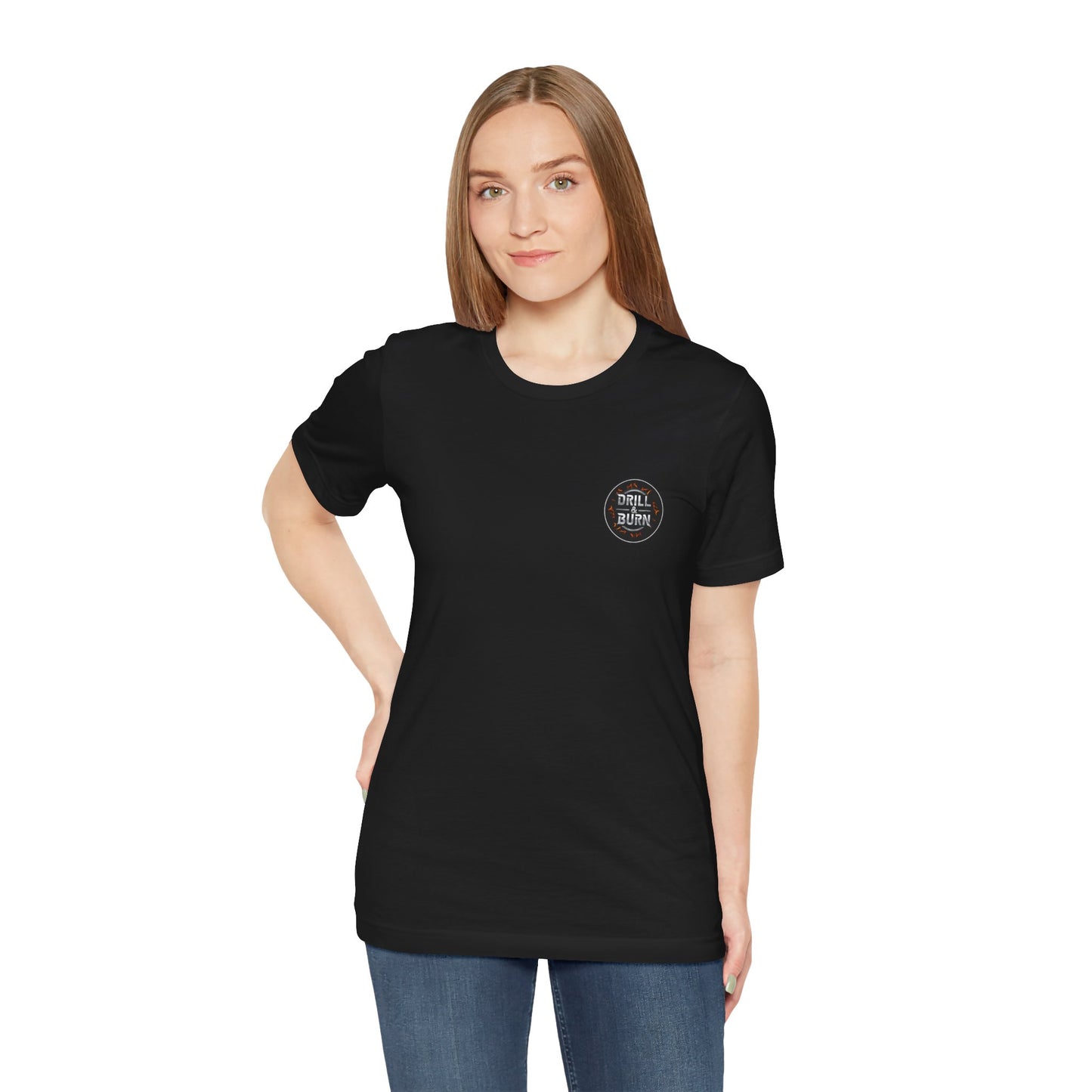 A woman with long brown hair, wearing a black Drill & Burn Graphic Unisex Jersey Tee from Printify and blue jeans, stands with one hand on her hip. The tee features soft Airlume cotton, a modern retail fit, and a small circular chest logo. She poses against a plain white background.