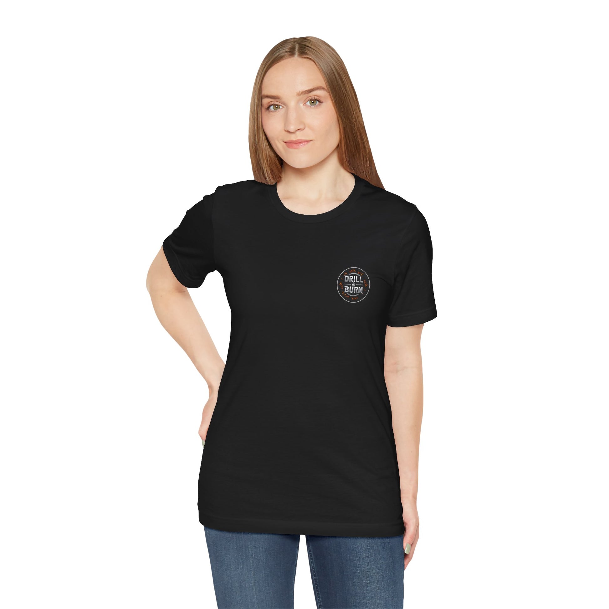A woman with long brown hair, wearing a black Drill & Burn Graphic Unisex Jersey Tee from Printify and blue jeans, stands with one hand on her hip. The tee features soft Airlume cotton, a modern retail fit, and a small circular chest logo. She poses against a plain white background.