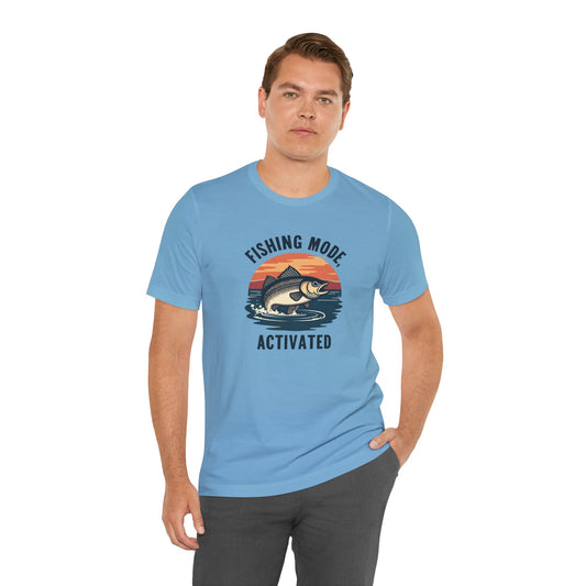 A person in a Printify Fishing Mode Activated Unisex Jersey Short Sleeve T-Shirt stands against a white background. The tee, crafted from soft Airlume cotton, features a light blue design with water and fish graphics. One hand is casually in their pocket.
