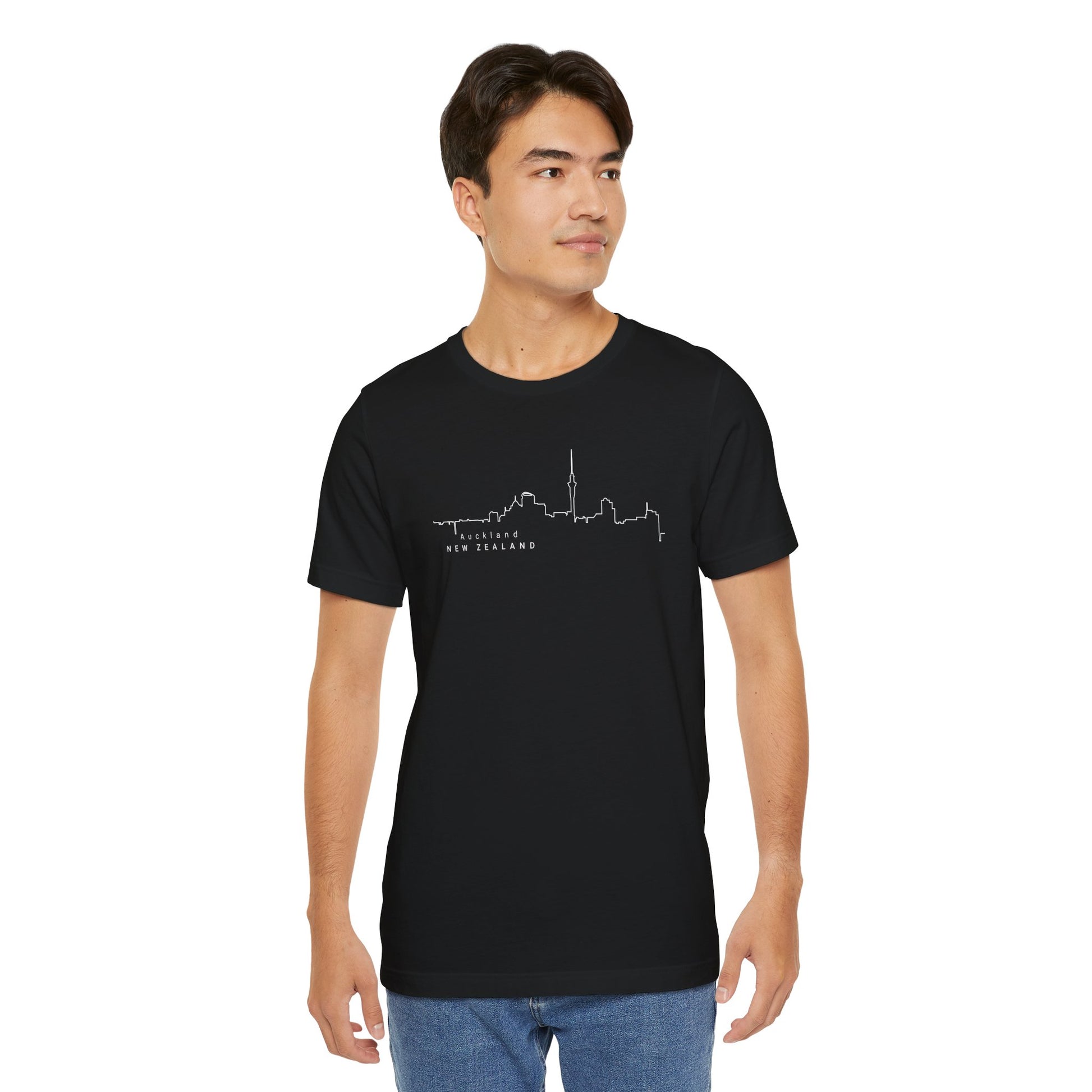 A person with short dark hair stands upright, looking sideways, and wears the Printifys Auckland Skyline Unisex Tee, a stylish black jersey featuring a white city skyline and New Zealand. Perfect for urban explorers, they pair it with light blue jeans.