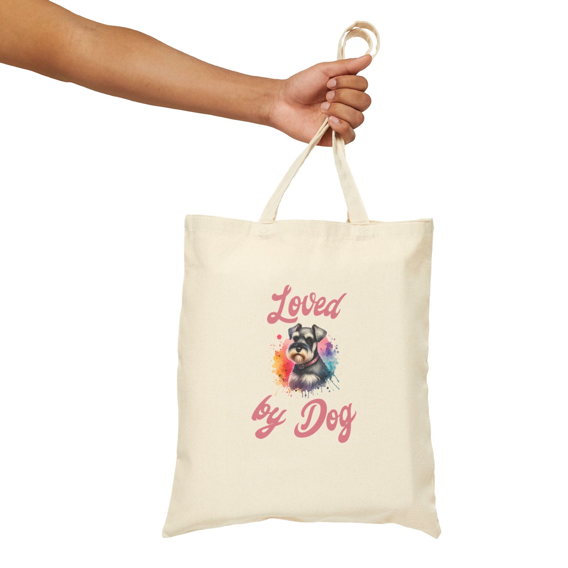 A hand holds the Loved By Dog Fun Cotton Canvas Tote Bag by D&B Apparel, made of 100% cotton, displaying Loved by Dog and a vibrant dogs face with bright paint splashes.