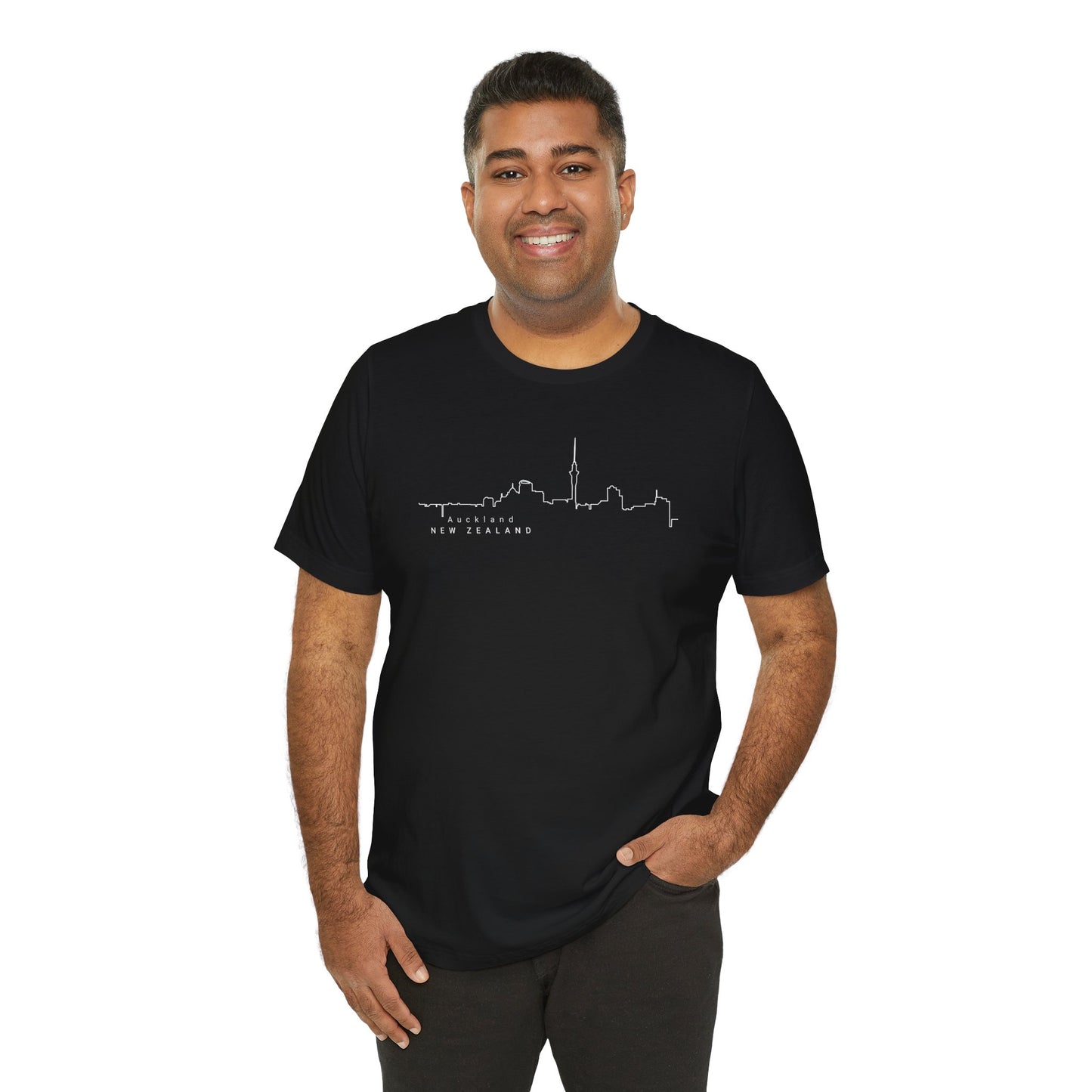 A smiling man with short dark hair stands against a white background wearing a black Auckland Skyline Unisex Tee by Printify. This sustainable jersey tee, perfect for travelers, features a white city skyline outline and New Zealand text. One hand rests in his pocket.