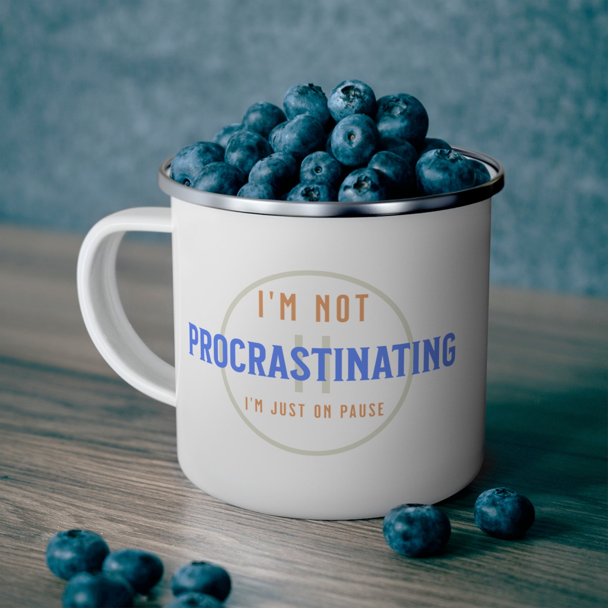 The Printify Funny Procrastination Enamel Camping Mug, ideal for outdoor lovers, sits on a wooden surface against a grayish-blue backdrop. It holds blueberries and features the text Im not procrastinating, Im just on pause, offering a perfect moment of stillness.