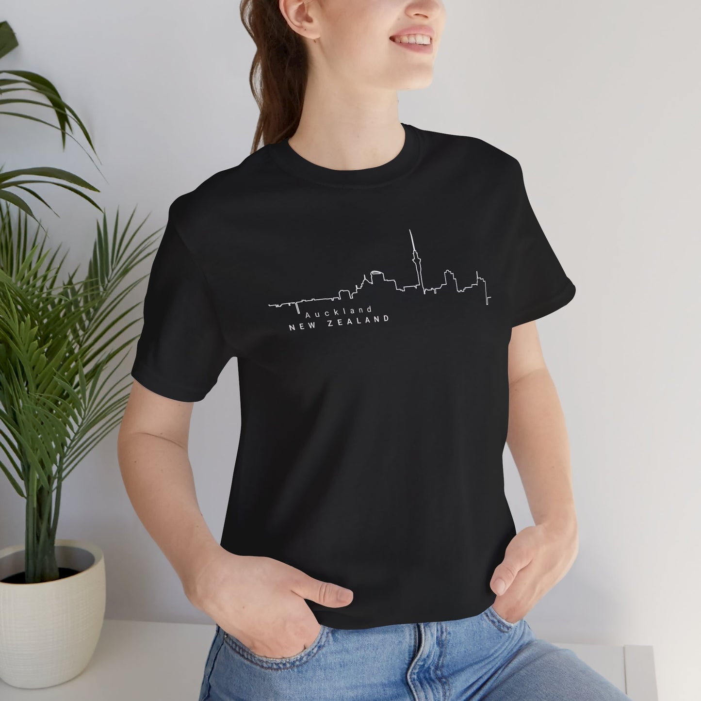 Wearing a Auckland Skyline Unisex Tee by Printify, this person sports a black jersey tee with a white cityscape outline and AUCKLAND NEW ZEALAND text.