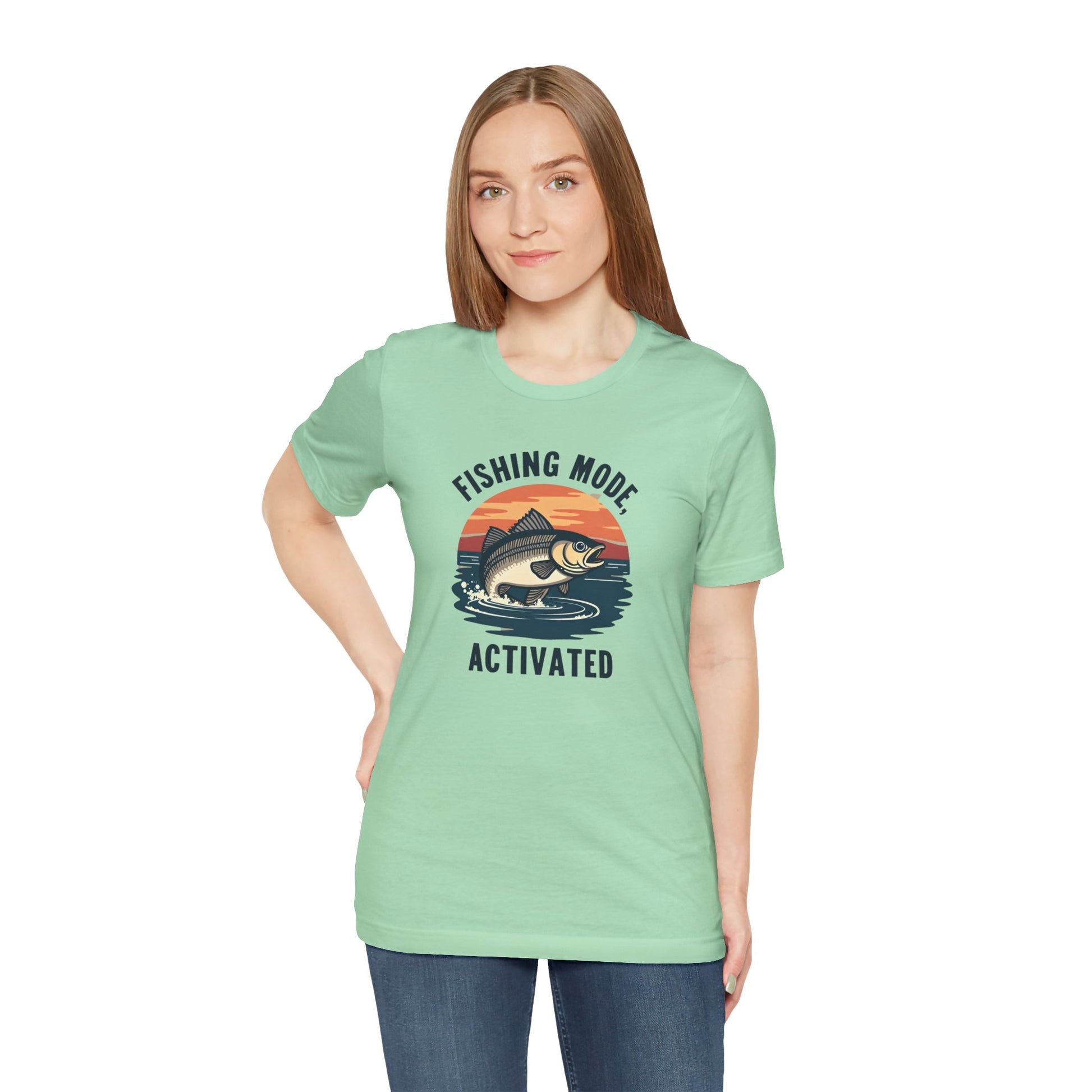 With long brown hair, they wear the Printify “Fishing Mode Activated” unisex short sleeve tee in light green, made from soft Airlume cotton. The shirt features a fish jumping against an orange sunset as they confidently pose with one hand on their hip.