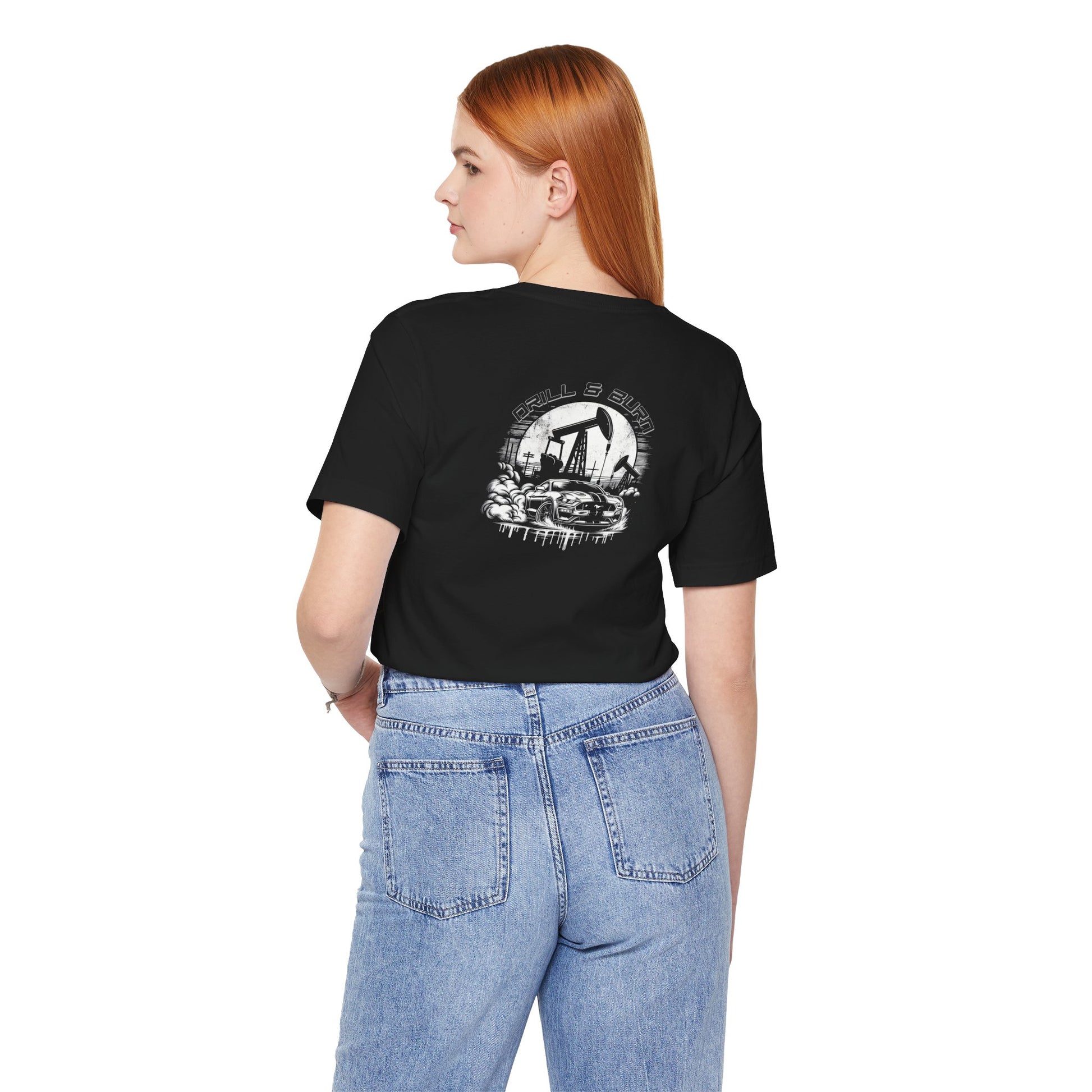 A woman with long red hair wears the Printify Drill & Burn Graphic Unisex Jersey Tee in black, featuring a skull and hand design on the back. She pairs it with light blue jeans made from soft Airlume cotton, standing with one hand in her back pocket.