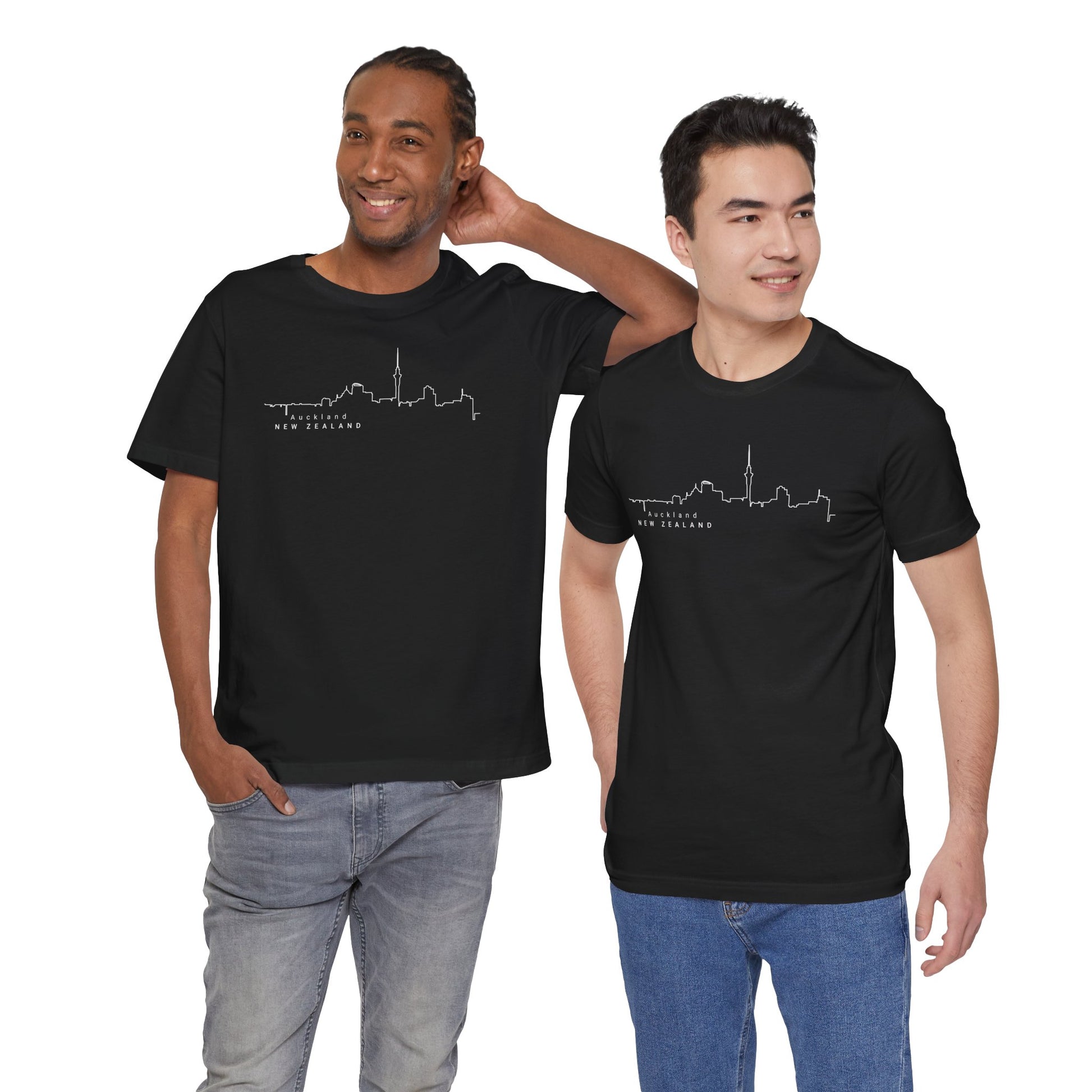 Two urban explorers in Printifys Auckland Skyline Unisex Tees, featuring a stylish white outline of a city skyline and New Zealand, stand together. One has a hand behind their head, both smiling against the plain white background, complemented by their casual pants.