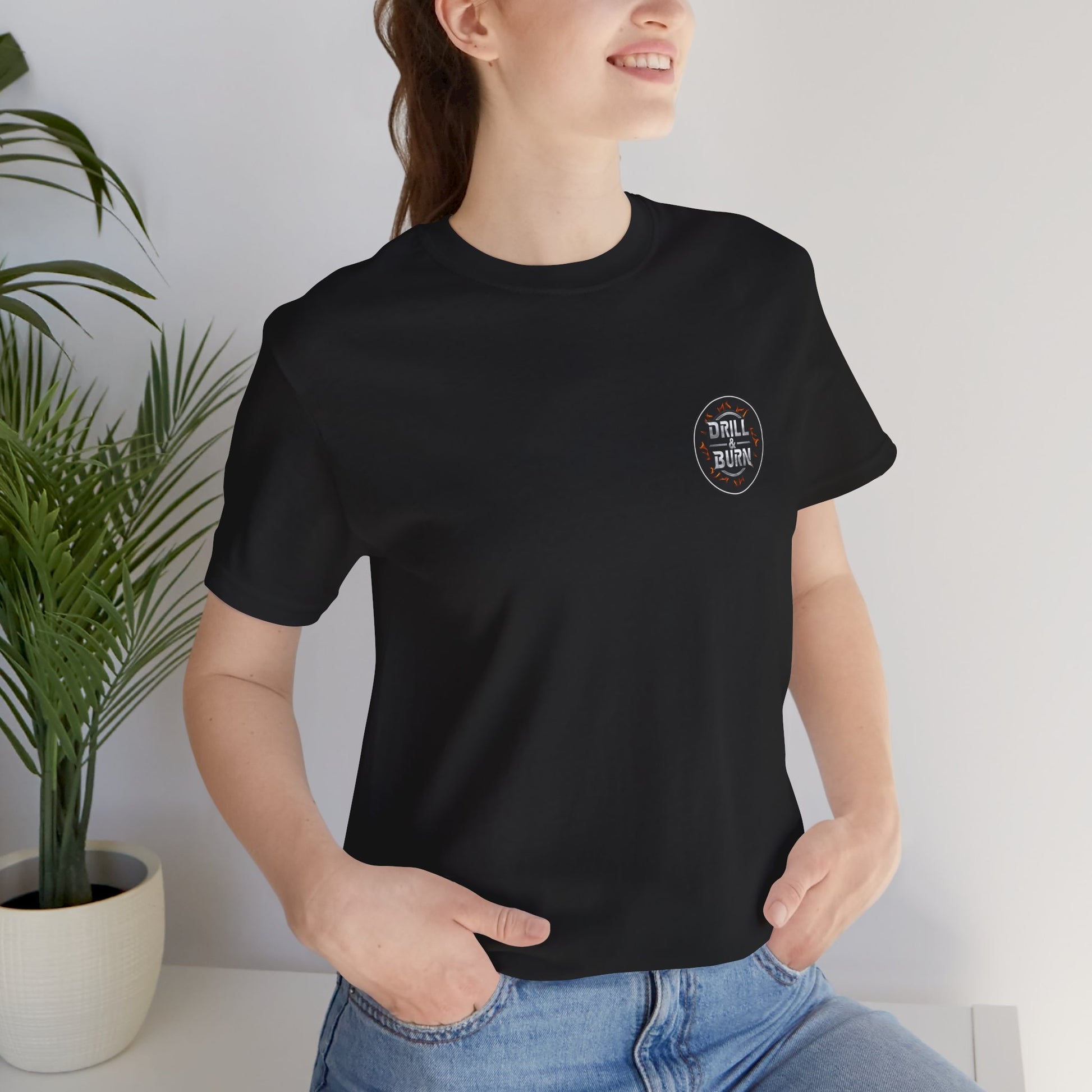 Wearing a black Drill & Burn Graphic Unisex Jersey Tee by Printify, featuring a small circular logo on the chest, a person smiles with hands in pockets. Crafted from Airlume cotton for a modern retail fit, they pose cheerfully next to a green plant in a white pot in the bright room.