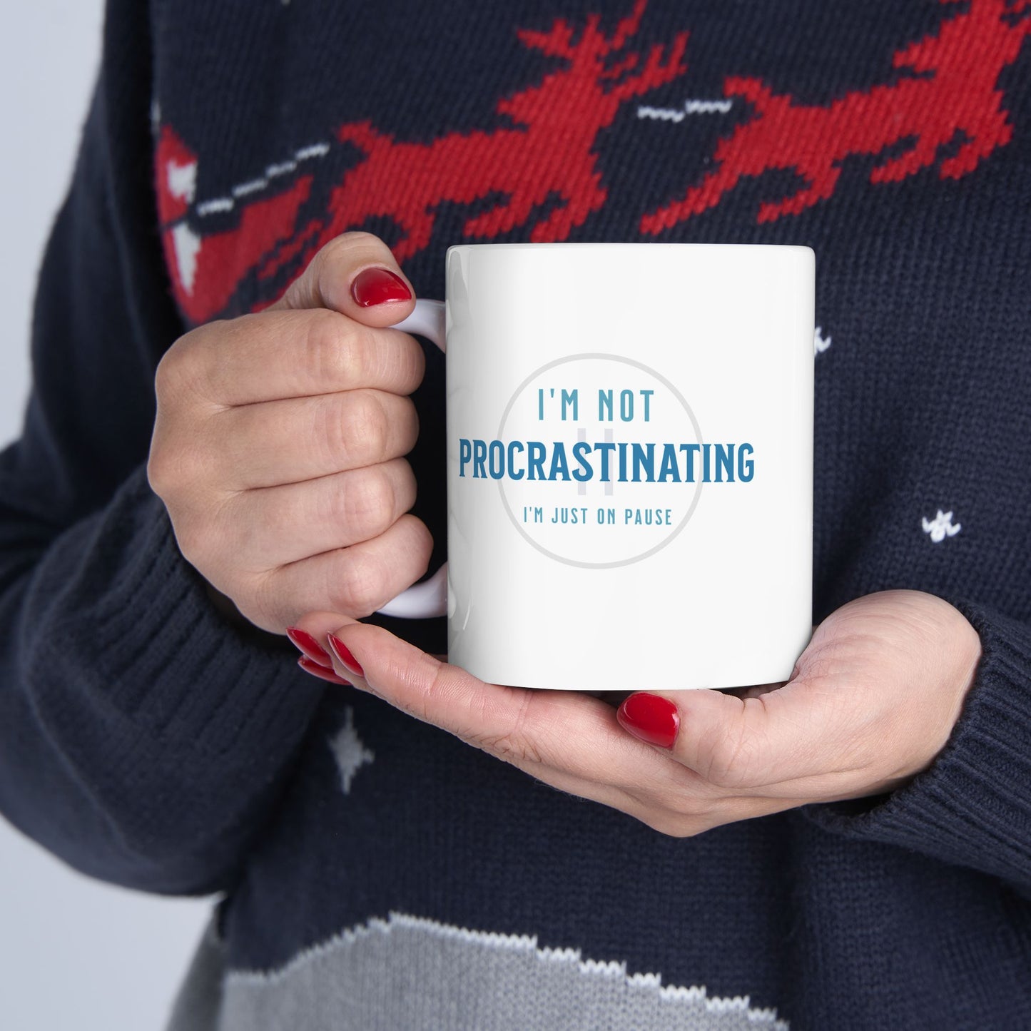 A person in a festive sweater holds a Printify Procrastination Ceramic Coffee Mug, featuring blue lettering that reads, Im not procrastinating, Im just on pause. The mug is ideal for coffee lovers and complements the persons red-painted nails and sweater design. Available in 11oz & 15oz.