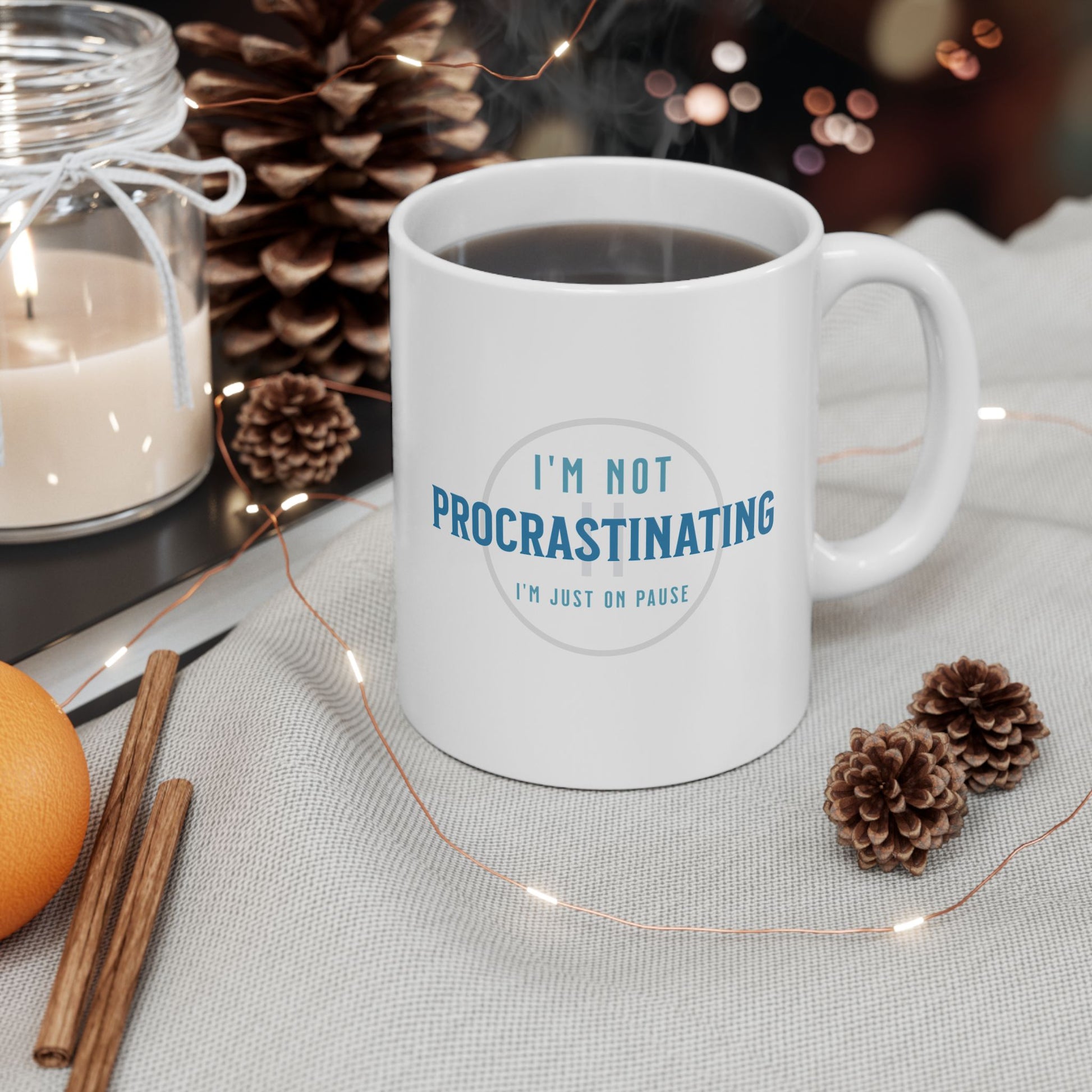 The Printify Procrastination Ceramic Coffee Mug rests on a gray cloth. Surrounded by a lit candle, pine cones, cinnamon sticks, an orange, and string lights, it creates a cozy atmosphere for coffee lovers.