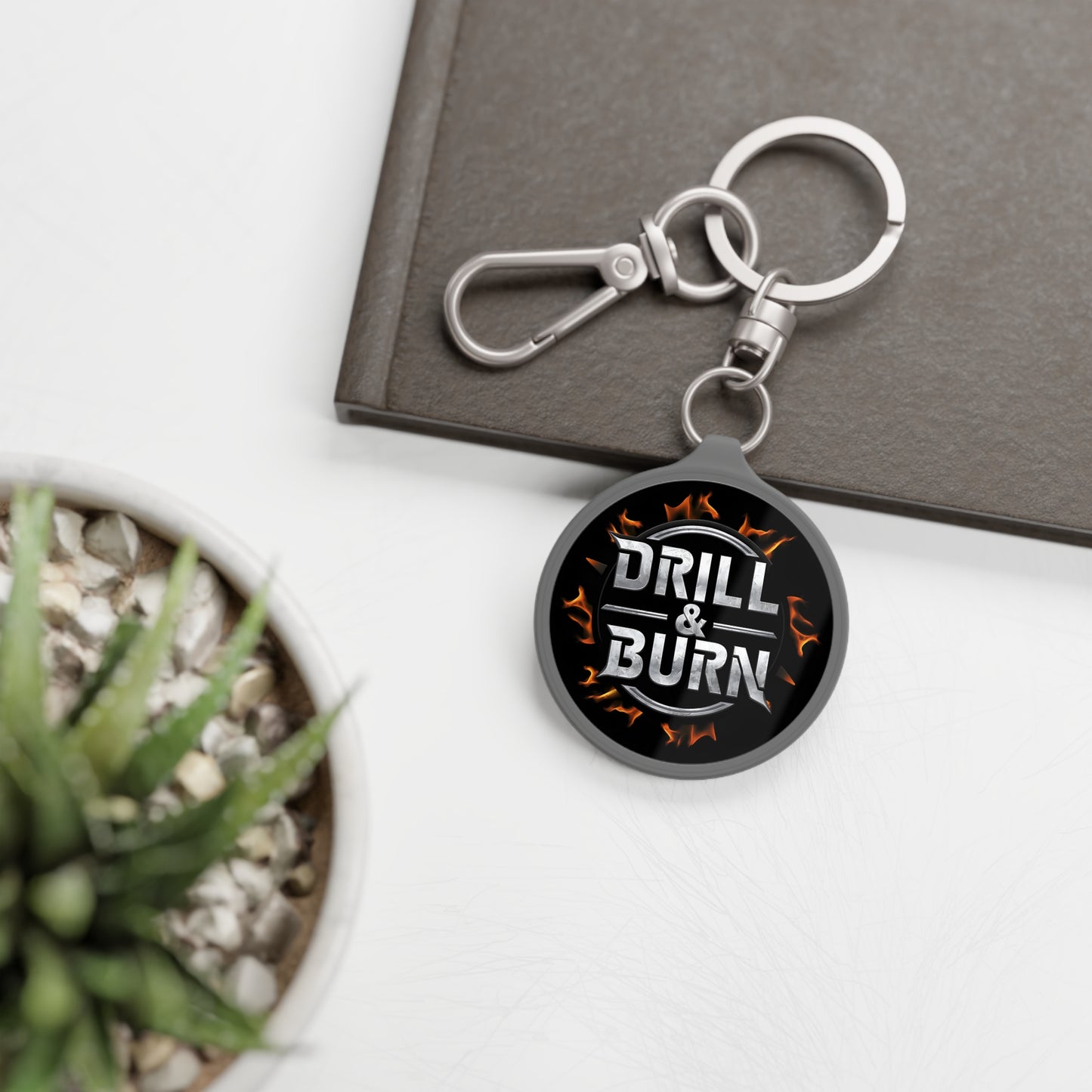 Acrylic keychain with a black DRILL & BURN tag and flame pattern on white from D&B Apparel. Perfect for personalized gifts, it pairs well with a succulent plant and gray notebook for car enthusiasts.