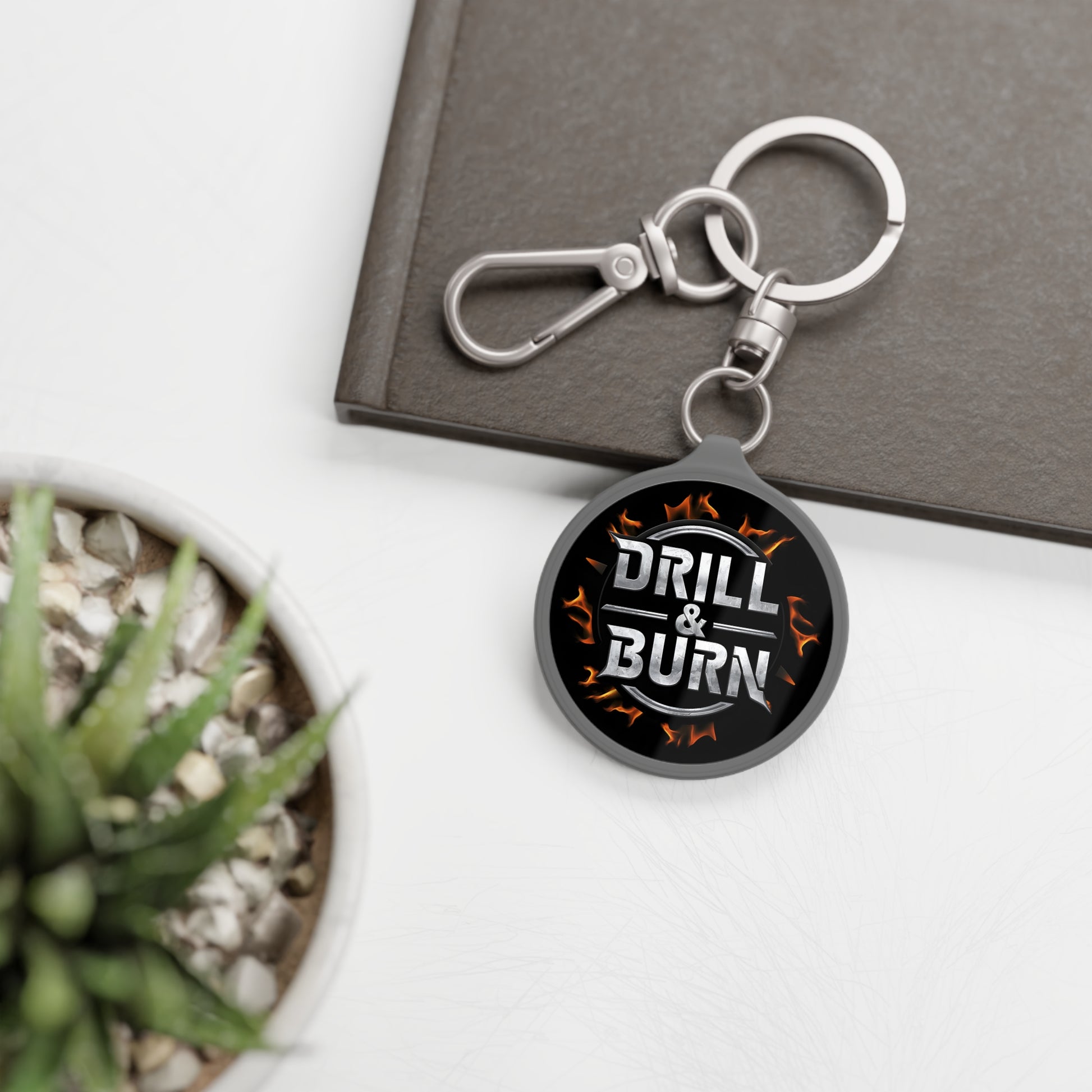 Acrylic keychain with a black DRILL & BURN tag and flame pattern on white from D&B Apparel. Perfect for personalized gifts, it pairs well with a succulent plant and gray notebook for car enthusiasts.