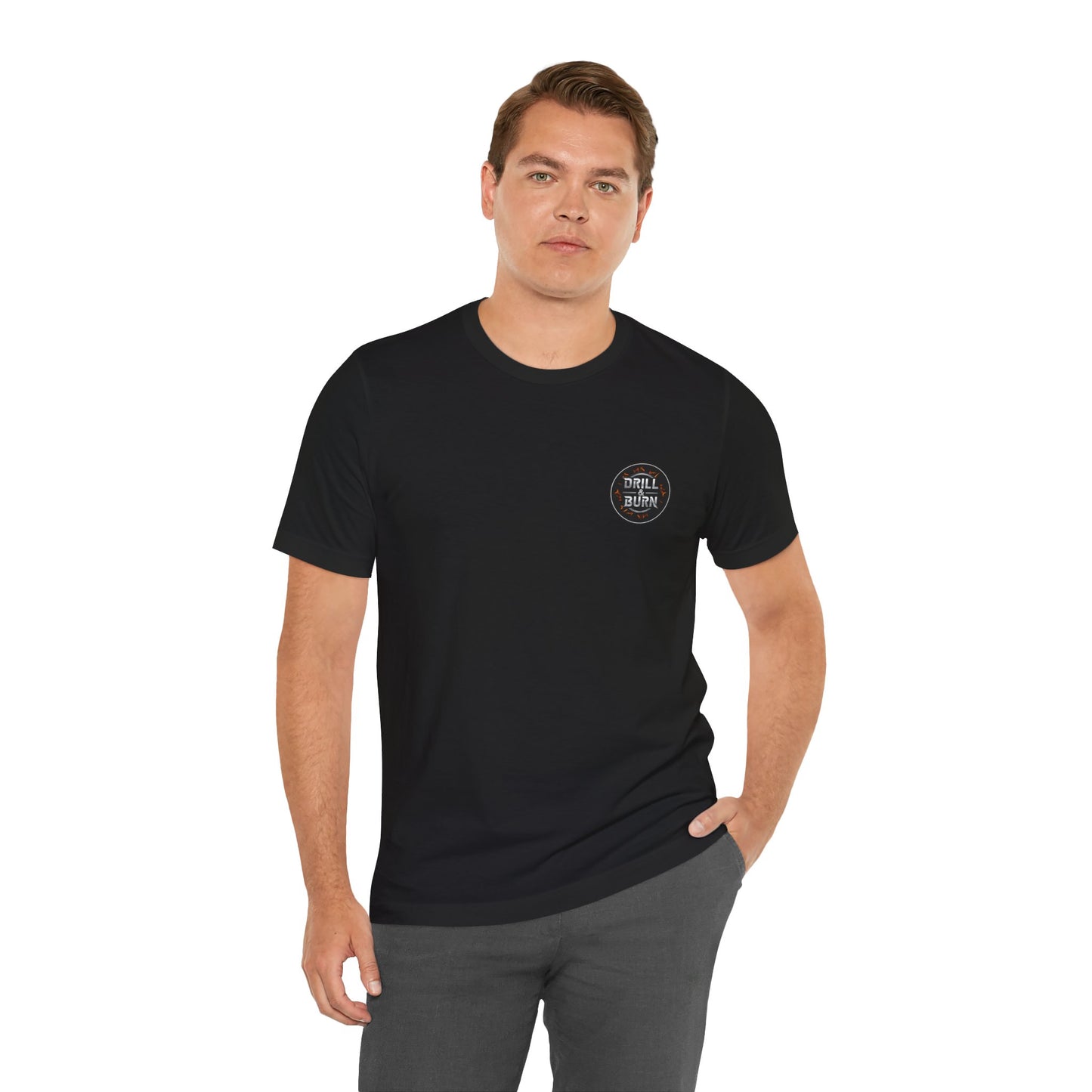 Standing against a white backdrop, a person in a black Drill & Burn Graphic Unisex Jersey Tee by Printify looks at the camera. Crafted from soft Airlume cotton with DISTILL KNIT on the chest and sporting short hair, they exude style in this modern retail fit, one hand in their pocket.