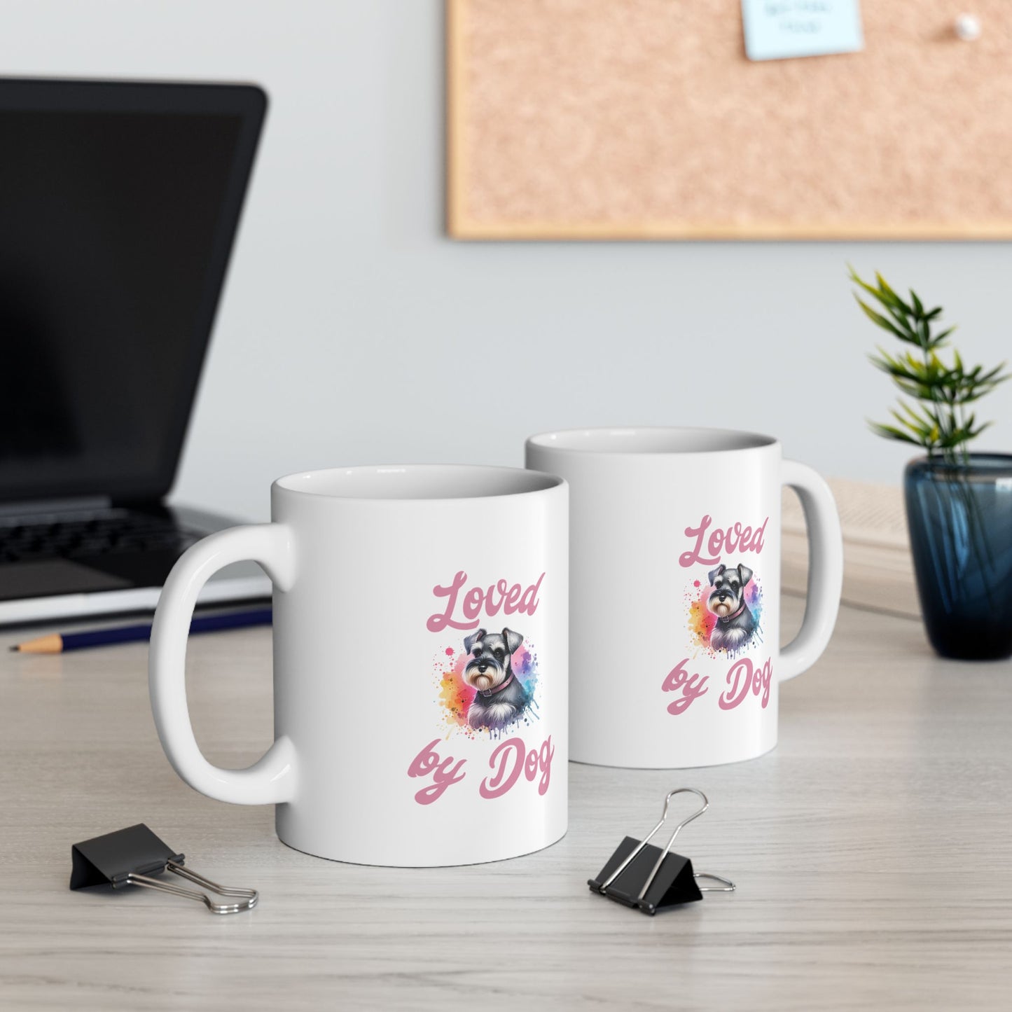 Two customized white ceramic mugs from D&B Apparel, each featuring the phrase Loved by Dog and an image of a miniature schnauzer, sit on a desk. Nearby are a laptop, plant, binder clips, and a corkboard with sticky notes in the background.