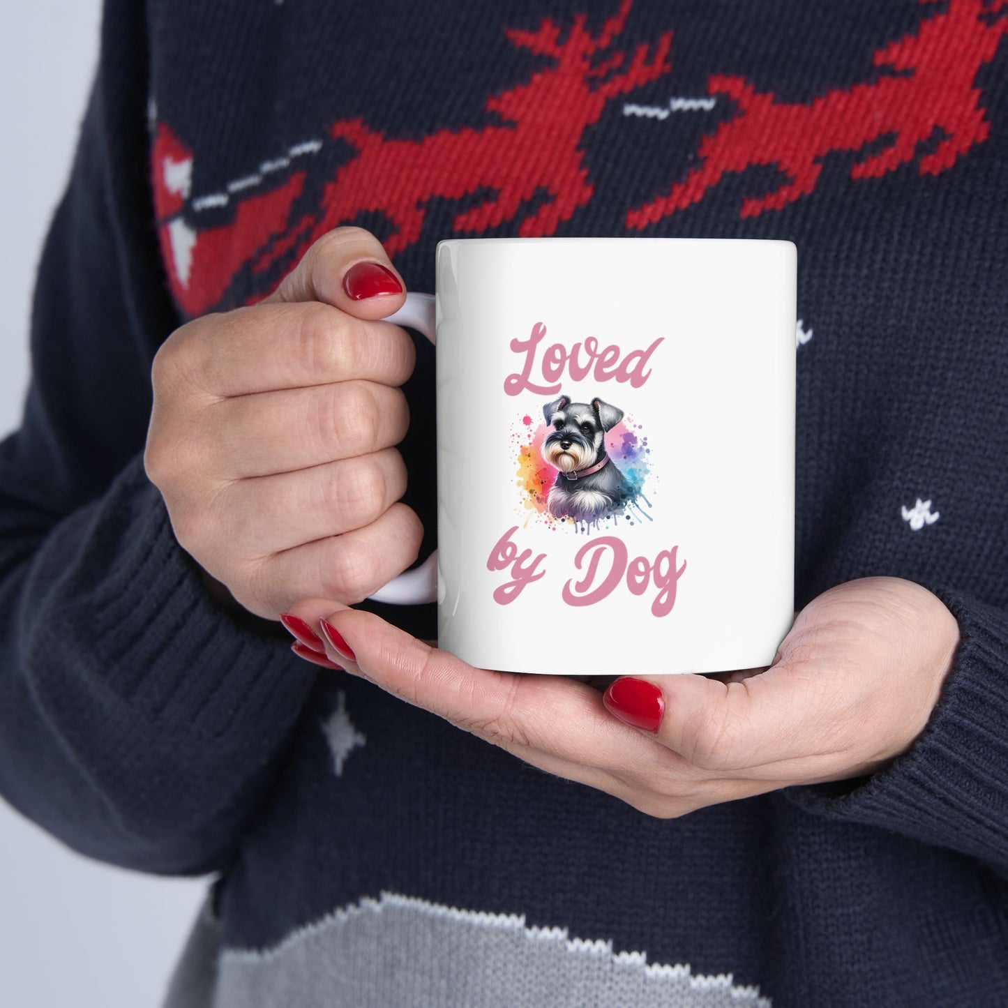 A person with red nail polish holds a D&B Apparel Miniature Schnauzer Ceramic Mug, customized with Loved by Dog and an image of a dog in sunglasses. This BPA-free mug complements their dark sweater featuring red deer patterns.