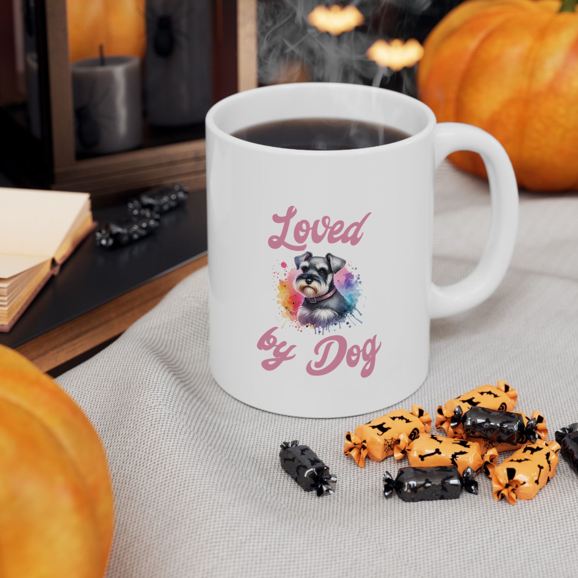A BPA-free ceramic mug from D&B Apparel, the Miniature Schnauzer Ceramic Mug (11oz, 15oz) displays Loved by Dog under a cartoon dog resembling a schnauzer. Filled with coffee, it sits among pumpkins, candles, and Halloween candies in black and orange wrappers.