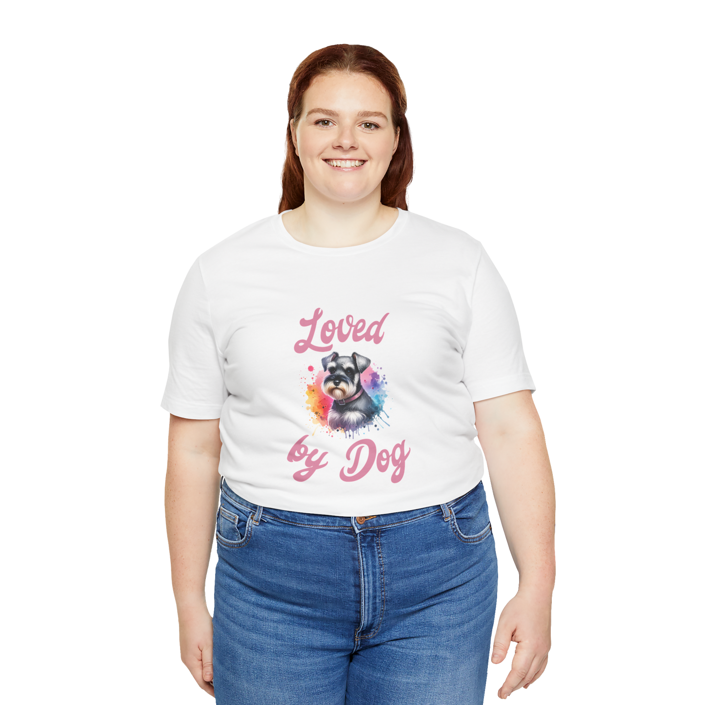 A red-haired person smiles in a white Loved by Dog t-shirt with a colorful miniature schnauzer, paired with blue jeans, standing against a black background. (D&B Apparel).