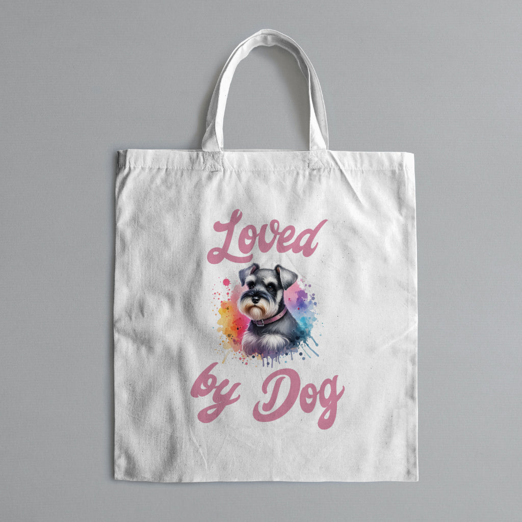 A ceramic mug features a miniature schnauzer with a collar against a light gray background. The design includes pink text stating Loved by Dog in watercolor style, available in 11oz and 15oz sizes from D&B Apparel.