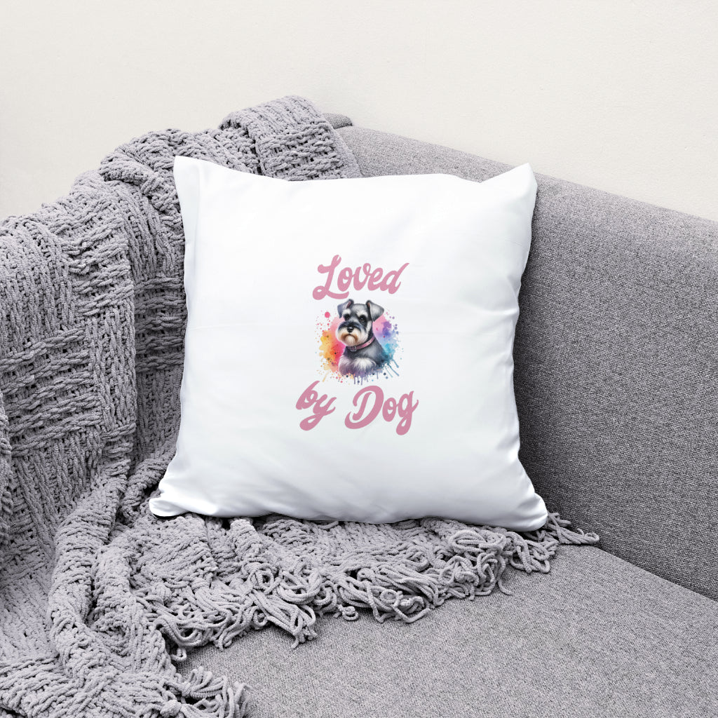 A Ceramic Mug from D&B Apparel features a vibrant Miniature Schnauzer design with the words Loved by Dog in pink, displayed on a gray couch beside a gray woven blanket and a decorative white pillow.
