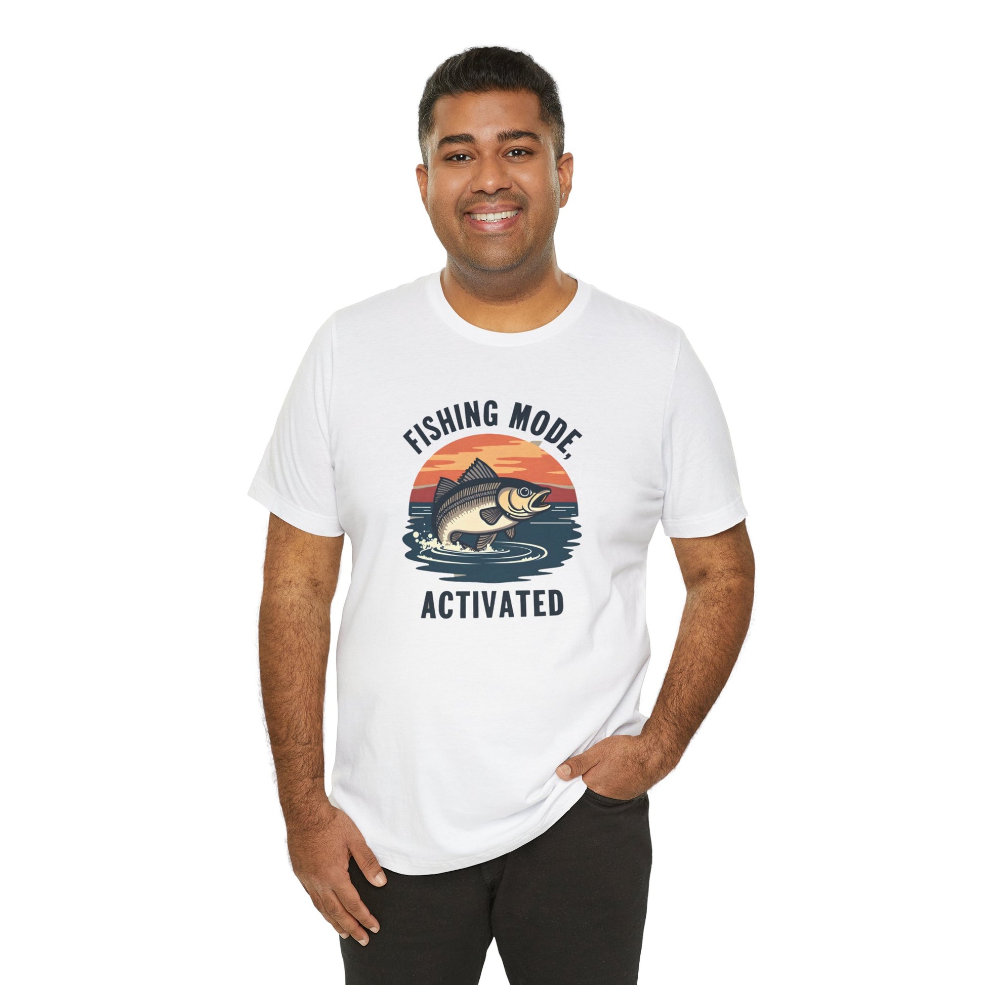 A person grins, wearing Printifys Fishing Mode Activated Tee for Anglers, a unisex white jersey short sleeve t-shirt made from soft Airlume cotton. It showcases a fish leaping at sunset with the caption Fishing Mode Activated. One hand rests casually in their pocket.