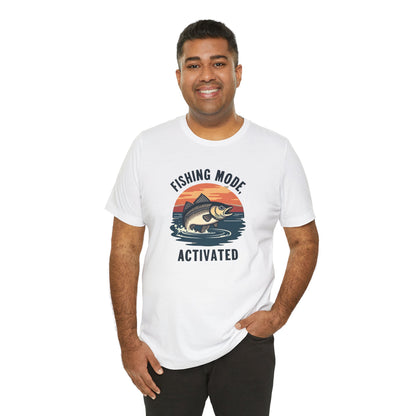 A person grins, wearing Printifys Fishing Mode Activated Tee for Anglers, a unisex white jersey short sleeve t-shirt made from soft Airlume cotton. It showcases a fish leaping at sunset with the caption Fishing Mode Activated. One hand rests casually in their pocket.
