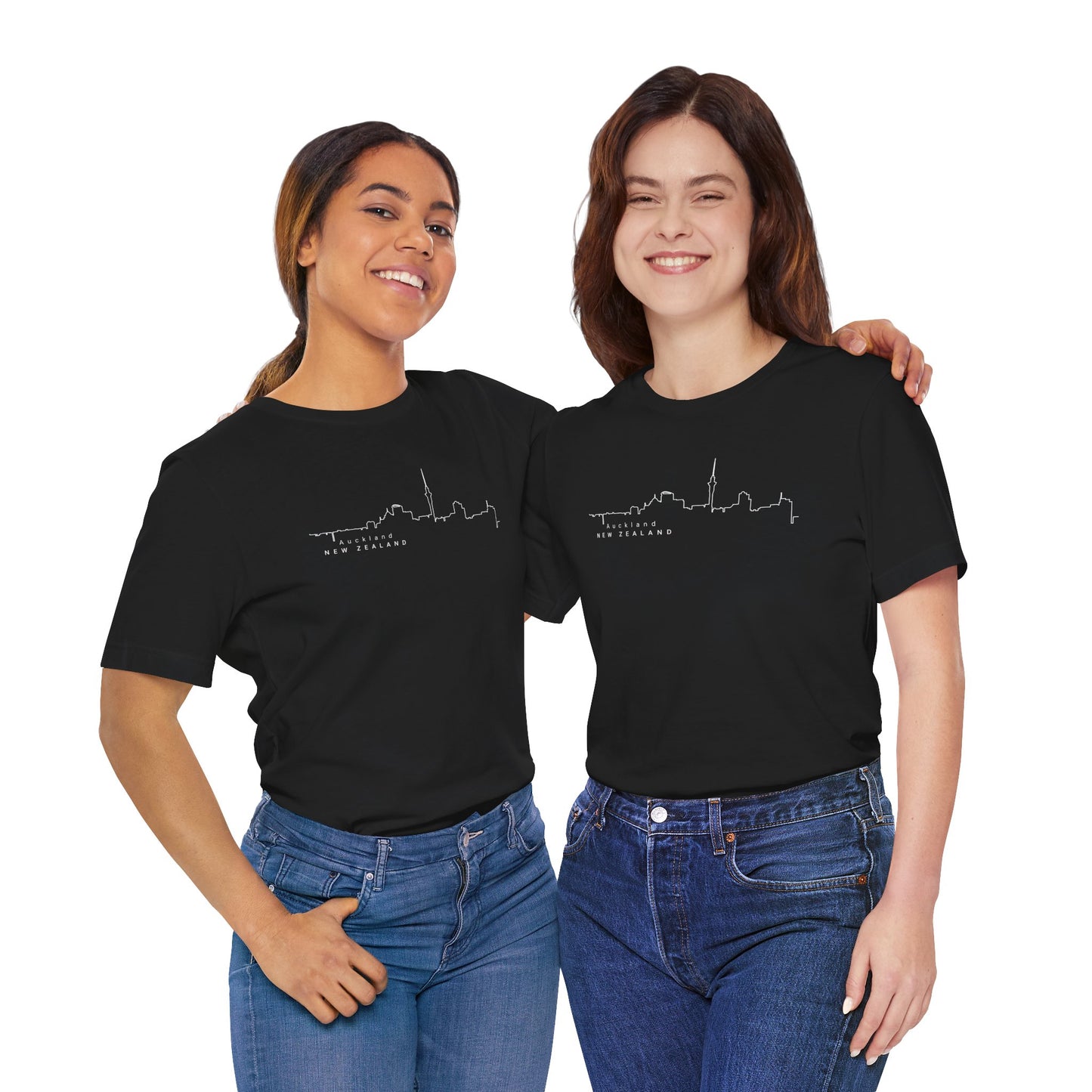 Two urban explorers stand together, smiling with their arms around each other, wearing Printifys Auckland Skyline Unisex Tee in black, crafted from sustainable material, and paired with blue jeans against a plain white background.