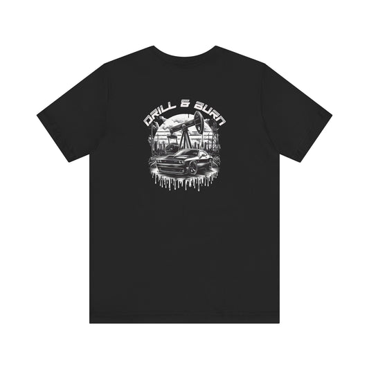 Black Drill & Burn Graphic Unisex Jersey Tee is shown from the front, highlighting an oil drilling scene with a Dodge Challenger doing a burnout. 